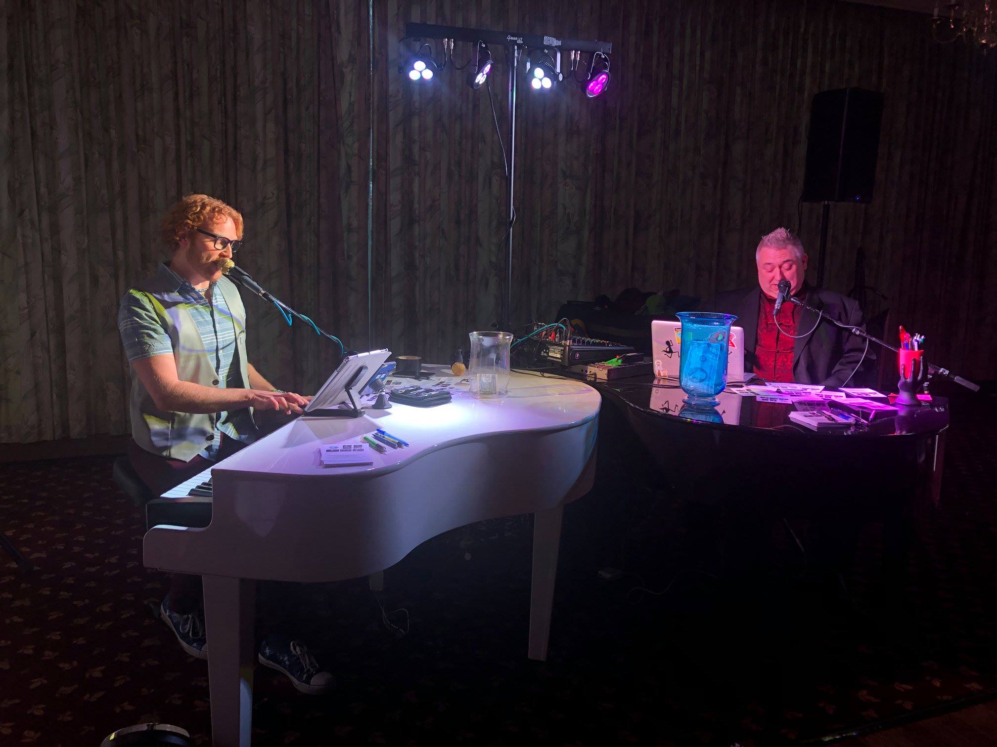 Two musicians playing pianos with microphones in a dimly lit room.