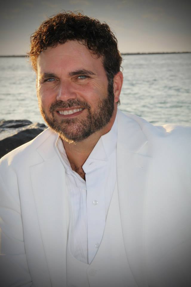 A man wearing a white suit smiles by the sea at sunset.