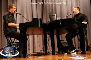 Two men performing a dueling piano show on stage with microphones.