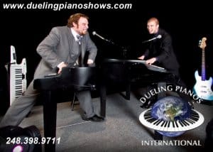 Two men performing at dueling pianos with guitars and keyboards.