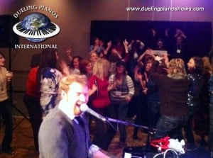 A performer sings at a lively Dueling Pianos event with an energetic crowd dancing.